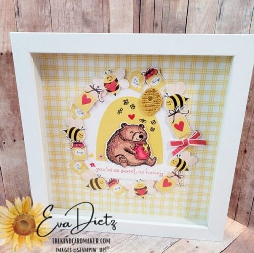 An 8 inch by 8 inch white framed home decor piece that shows a bear with a pot of honey sitting in the middle, surrounded by bumblebees and jars of honey. One red bow finishes it off. Designed by Eva Dietz, The Kind Card Maker.